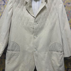 May include: A light beige corduroy blazer with a quilted diamond pattern. The blazer has three buttons, two front pockets, and a notched lapel. The jacket is hanging on a grey hanger.