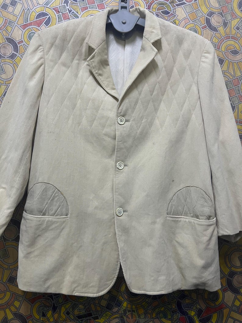 May include: A light beige corduroy blazer with a quilted diamond pattern. The blazer has three buttons, two front pockets, and a notched lapel. The jacket is hanging on a grey hanger.