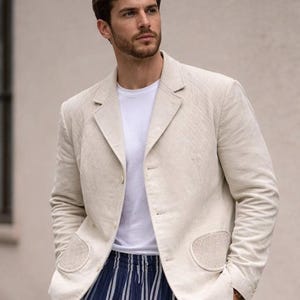 May include: A light beige blazer with a quilted design, worn with a white t-shirt and blue and white striped trousers. The blazer features two front pockets and a notched lapel. The ensemble is casual and fashionable.
