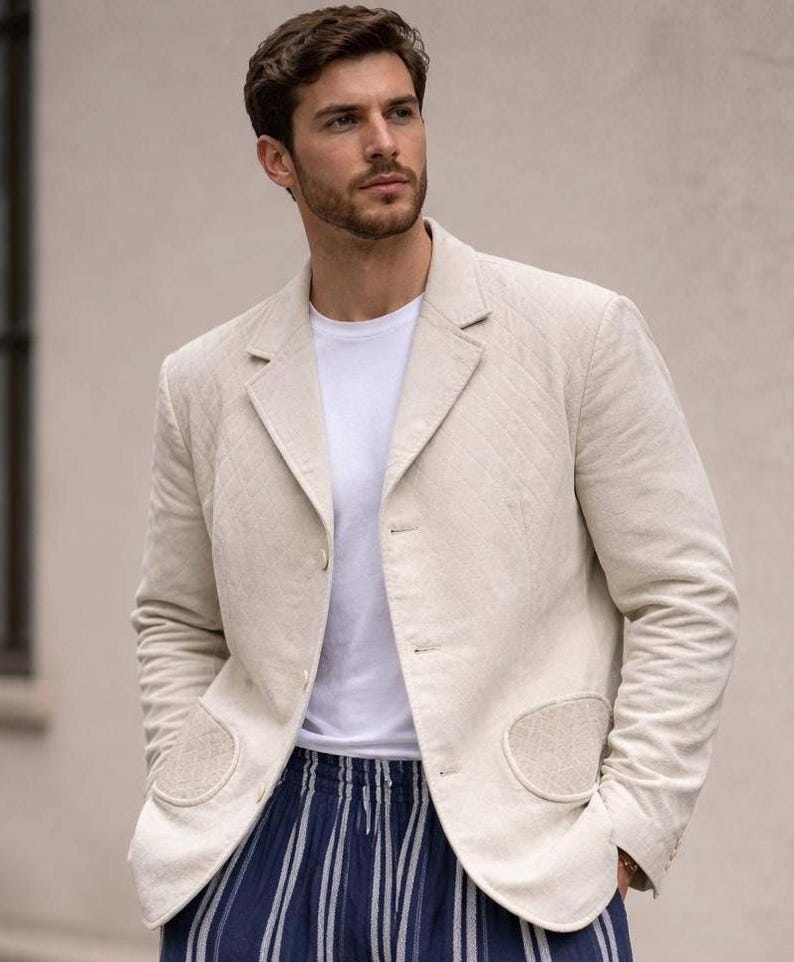 May include: A light beige blazer with a quilted design, worn with a white t-shirt and blue and white striped trousers. The blazer features two front pockets and a notched lapel. The ensemble is casual and fashionable.