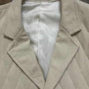May include: A beige corduroy blazer with a quilted diamond pattern. The blazer features a notched lapel and a white lining. The fabric has a ribbed texture. The blazer is a neutral colour and suitable for anyone.