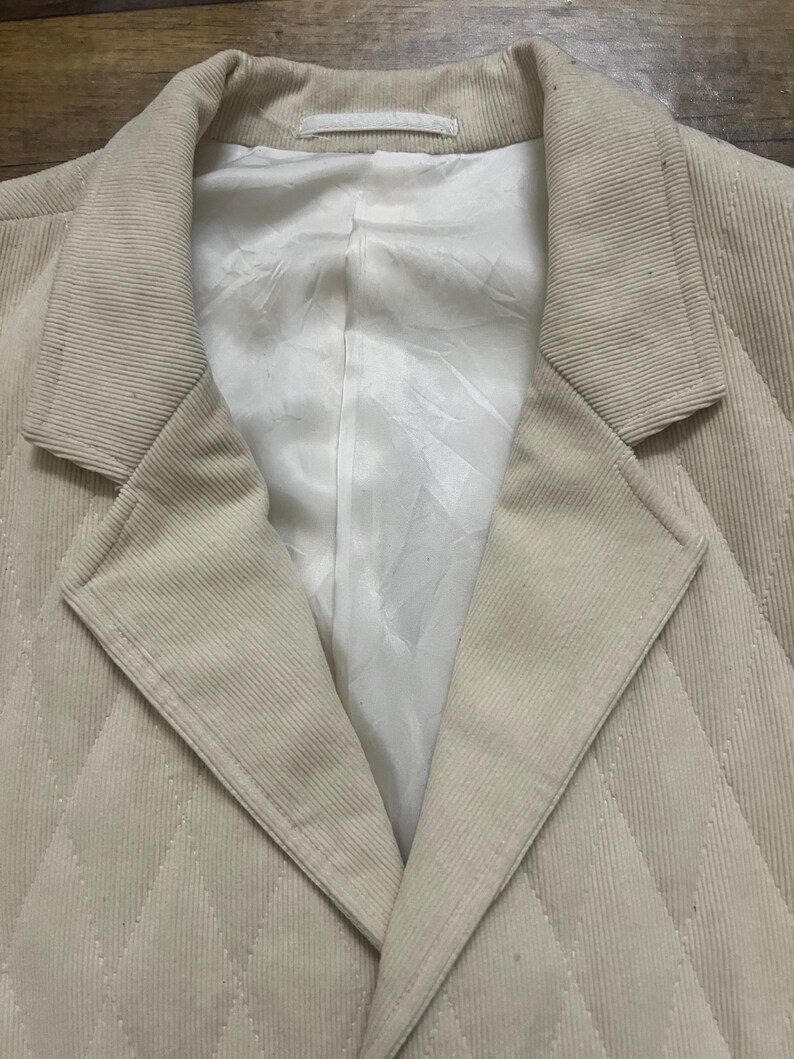 May include: A beige corduroy blazer with a quilted diamond pattern. The blazer features a notched lapel and a white lining. The fabric has a ribbed texture. The blazer is a neutral colour and suitable for anyone.