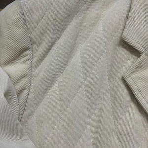 May include: Close-up of a beige corduroy blazer. The jacket has a quilted diamond pattern on the front and a classic notched lapel. The fabric has a ribbed texture, and the stitching is visible.
