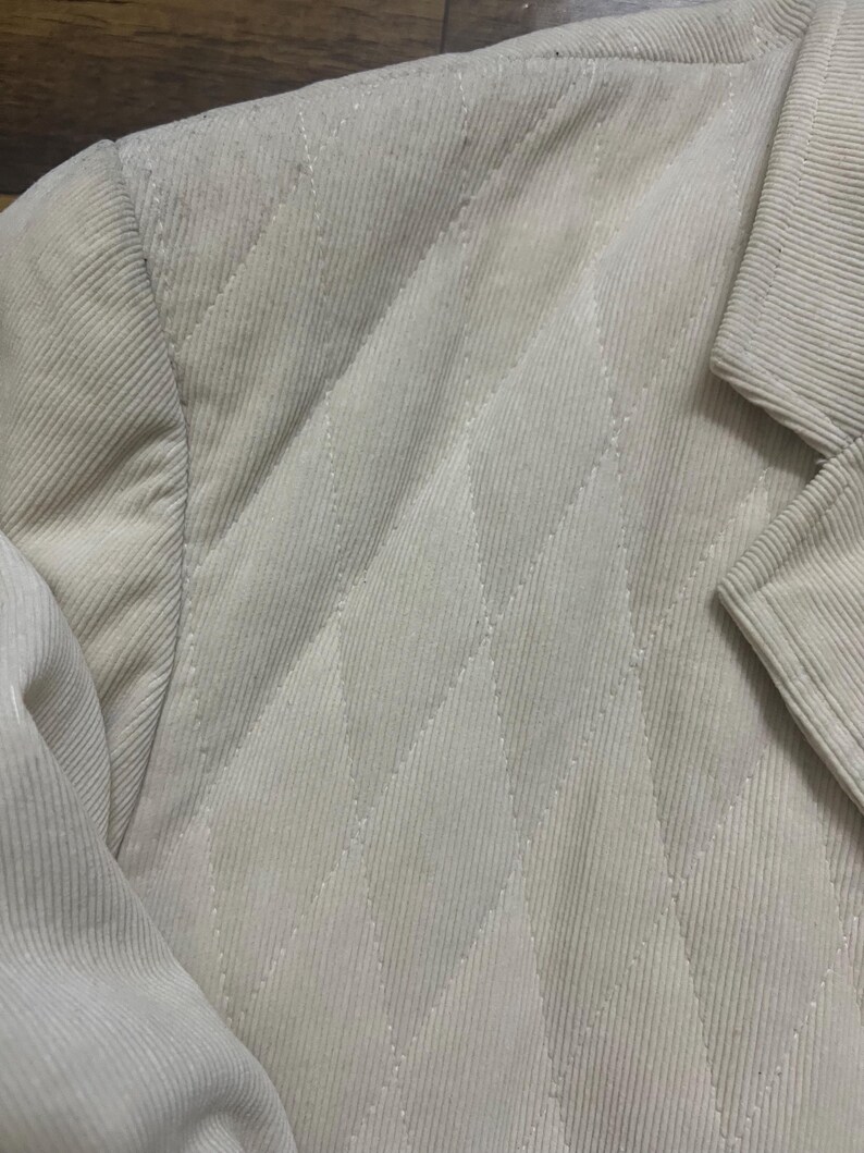 May include: Close-up of a beige corduroy blazer. The jacket has a quilted diamond pattern on the front and a classic notched lapel. The fabric has a ribbed texture, and the stitching is visible.