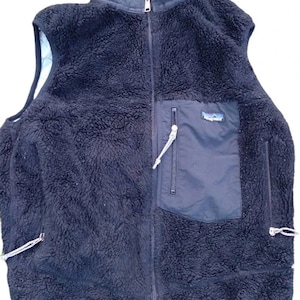 Könnte beinhalten: Eine schwarze Fleece-Weste mit hohem Kragen und durchgehendem Reißverschluss. Die Weste hat eine dunkelblaue Brusttasche mit Reißverschluss und ein kleines Markenlogo. Weiße Kordelzüge sind an den Taschen sichtbar.