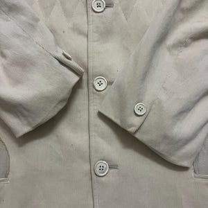 May include: A close-up shot of a light beige corduroy blazer. The jacket has a diamond-patterned design on the upper section and is fastened with three white buttons. The sleeves have buttoned cuffs, and the style is classic.