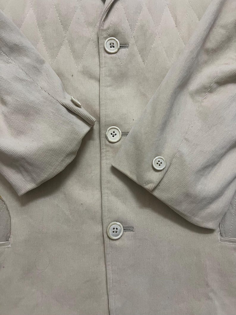 May include: A close-up shot of a light beige corduroy blazer. The jacket has a diamond-patterned design on the upper section and is fastened with three white buttons. The sleeves have buttoned cuffs, and the style is classic.