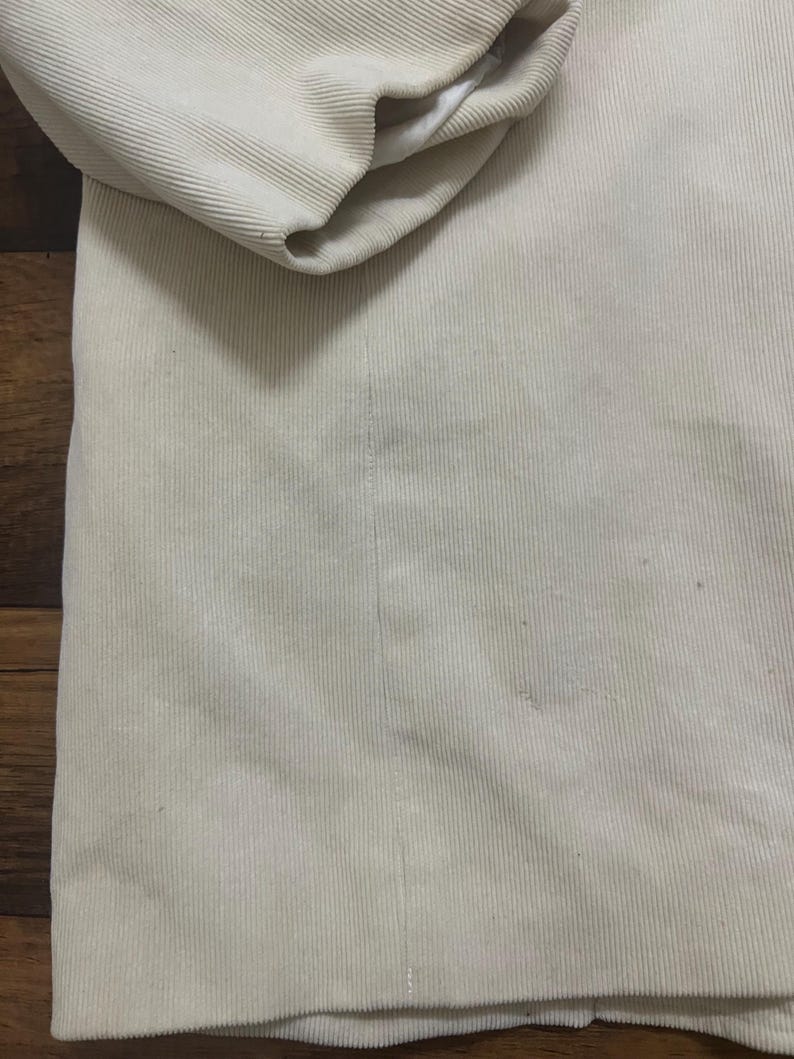May include: A close-up view of a beige corduroy jacket. The jacket has a ribbed texture and a folded sleeve, showing a white lining. The fabric appears soft and has a slight sheen. The jacket is a neutral colour.