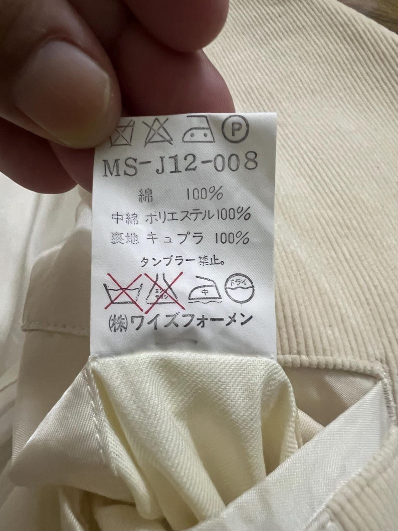 May include: A close-up of a cream-coloured fabric tag with Japanese text and care symbols. The tag reads "MS-J12-008" and lists fabric composition: 100% cotton, 100% polyester, and 100% cupro.