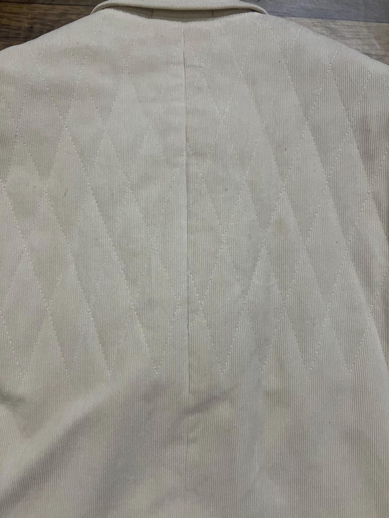 May include: Cream-coloured blazer featuring a diamond pattern. The jacket has a classic collar and a vertical seam at the back. The fabric has a corduroy texture.