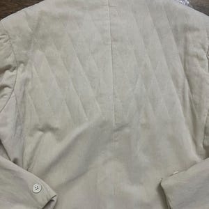 May include: A beige blazer with a diamond-quilted back. The blazer features a buttoned sleeve and a traditional collar. The fabric has a ribbed texture. The garment is likely made of corduroy or a similar material.