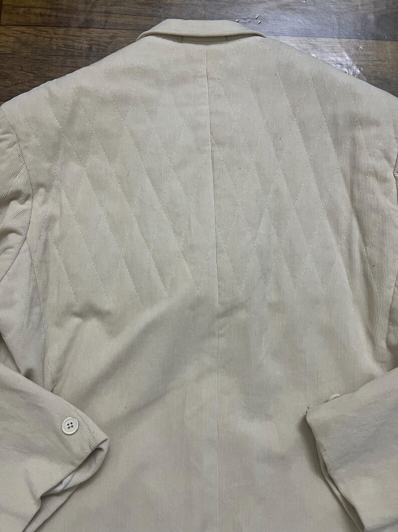 May include: A beige blazer with a diamond-quilted back. The blazer features a buttoned sleeve and a traditional collar. The fabric has a ribbed texture. The garment is likely made of corduroy or a similar material.