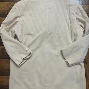 May include: A cream-coloured blazer with a quilted back design. The blazer has long sleeves, a notched lapel, and two buttons on the front. The back features a diamond pattern. The blazer is made of a soft, textured fabric.
