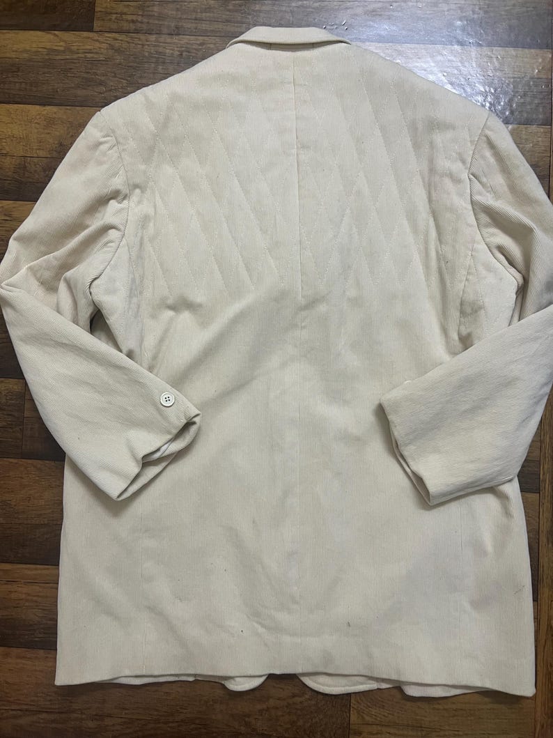 May include: A cream-coloured blazer with a quilted back design. The blazer has long sleeves, a notched lapel, and two buttons on the front. The back features a diamond pattern. The blazer is made of a soft, textured fabric.