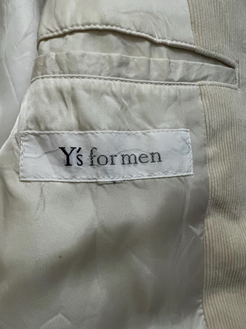 May include: Close-up of the interior of a light beige corduroy blazer. The lining is a lighter shade of beige. A white tag with the text "Y's for men" is sewn into the lining.