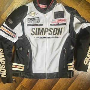 Może przedstawiać: Czarna, biała i szara kurtka motocyklowa Simpson z wieloma naszywkami i logo Simpson na piersi. Kurtka ma zapięcie na zamek błyskawiczny, czarne skórzane akcenty i napis "The Winning Edge".