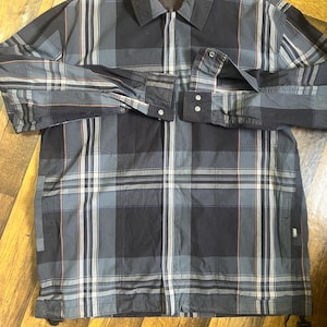 以下が含まれることがあります： 黒、グレー、青のチェック柄ジャケット。フルレングスのジッパーとボタン付きの袖口が付いています。 ジャケットは襟付きで、裾にドローコードが付いています。 チェック柄には、細い赤と白の線が含まれています。