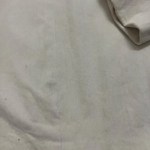May include: Close-up of cream-coloured corduroy fabric. The fabric has vertical ridges and a soft texture. A folded sleeve is visible in the upper right corner. The fabric appears to be part of a garment.