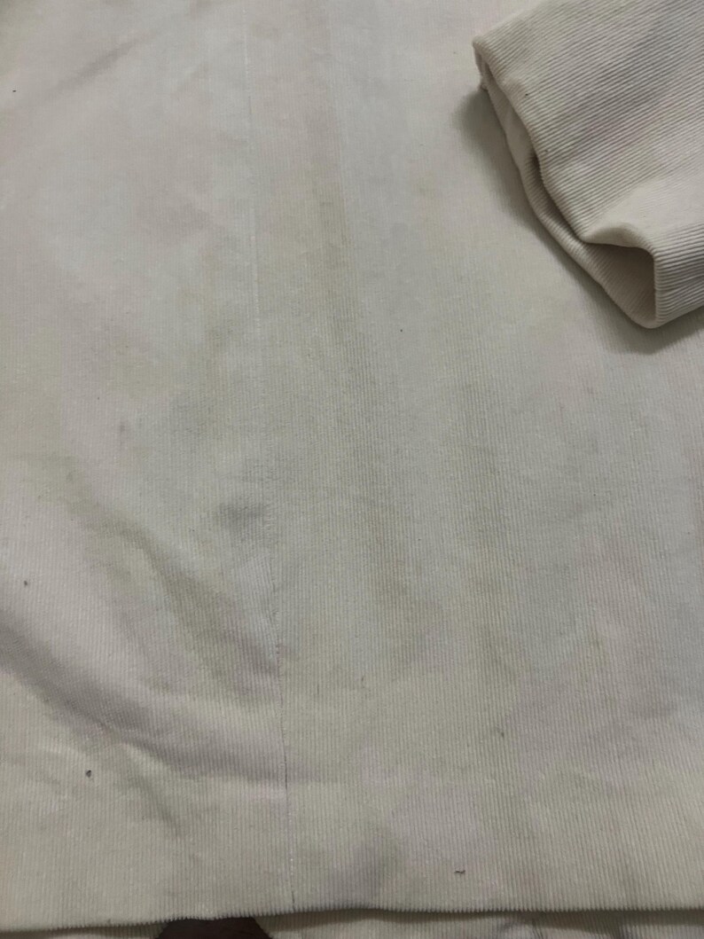 May include: Close-up of cream-coloured corduroy fabric. The fabric has vertical ridges and a soft texture. A folded sleeve is visible in the upper right corner. The fabric appears to be part of a garment.