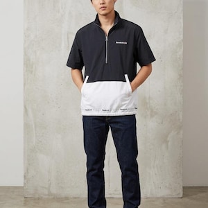 Op de afbeelding: Zwart-wit jack met korte mouwen en een ritssluiting, gecombineerd met een donkerblauwe jeans en witte sneakers. De jas heeft het Reebok-logo op de borst en een wit paneel aan de onderkant. De jeans heeft een rechte pijp.