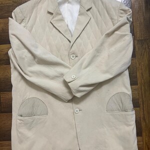 May include: A beige blazer with a quilted diamond pattern. It features two front pockets, a notched lapel, and three buttons. The interior lining is white. The jacket is a neutral colour.
