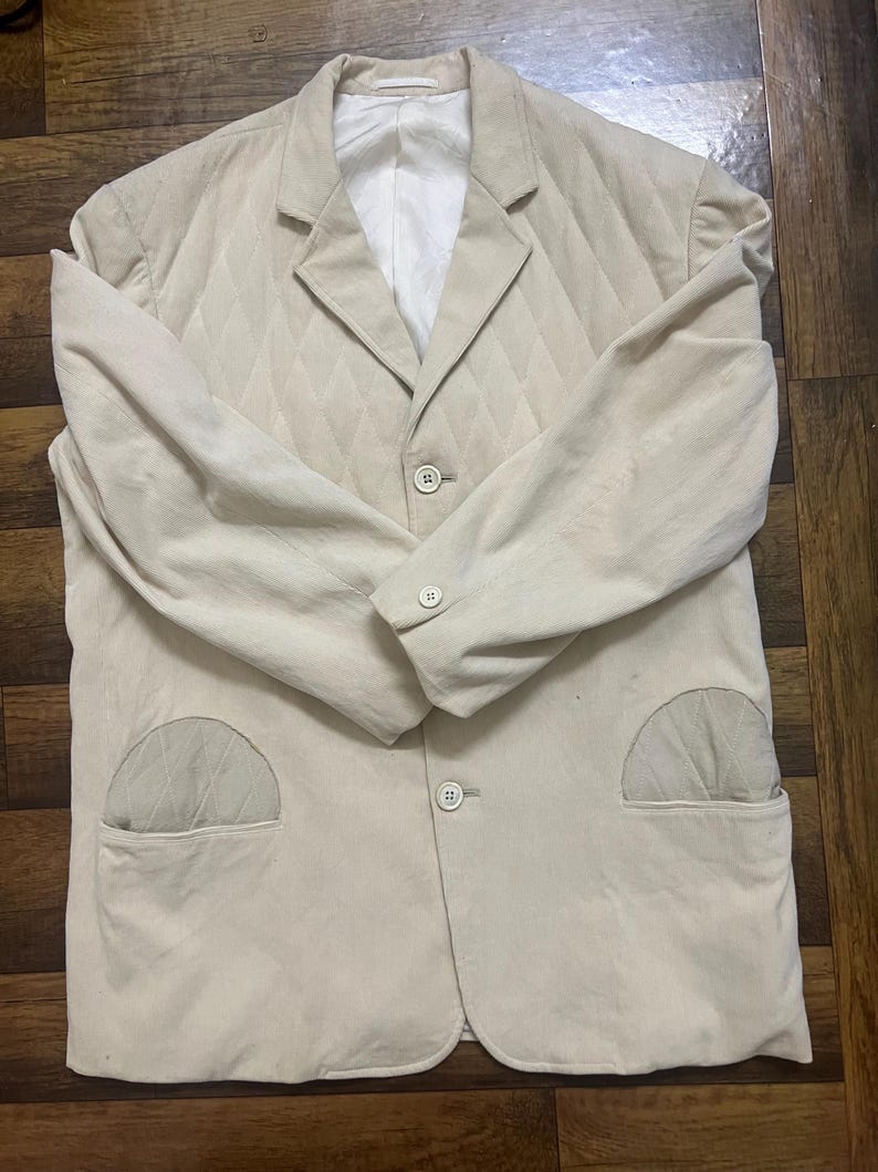May include: A beige blazer with a quilted diamond pattern. It features two front pockets, a notched lapel, and three buttons. The interior lining is white. The jacket is a neutral colour.