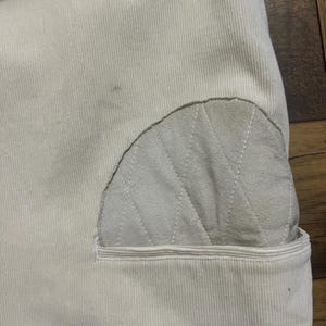 May include: Detail of cream corduroy fabric with a light grey, quilted patch. The patch features a diamond stitch pattern and is sewn onto a pocket. The fabric has vertical ridges.