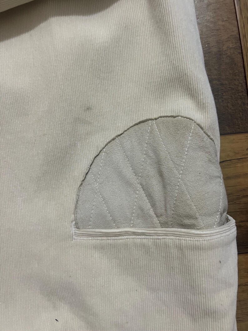 May include: Detail of cream corduroy fabric with a light grey, quilted patch. The patch features a diamond stitch pattern and is sewn onto a pocket. The fabric has vertical ridges.