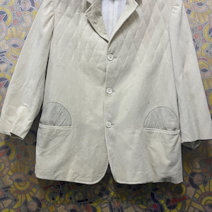 May include: A light beige blazer with a quilted diamond pattern. The blazer has three buttons, two front pockets, and a notched lapel. The jacket is hanging on a grey hanger.