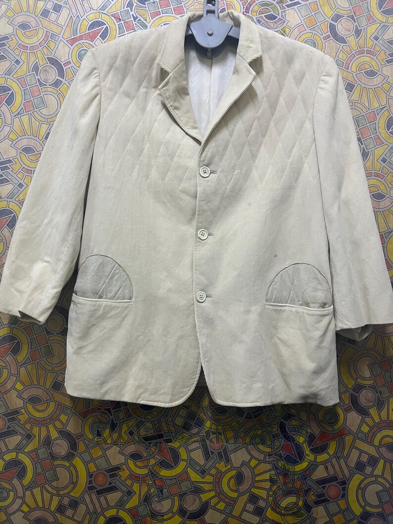 May include: A light beige blazer with a quilted diamond pattern. The blazer has three buttons, two front pockets, and a notched lapel. The jacket is hanging on a grey hanger.