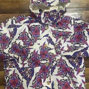 May include: A white hooded jacket with a vibrant pattern of purple, red, and blue abstract shapes. The jacket features a button-up front, pockets, and gold-colored buttons. The jacket is a vintage style.