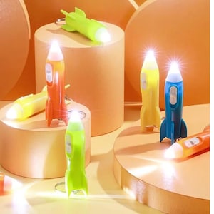 Może przedstawiać: Zestaw sześciu kolorowych latarek w kształcie rakiety, które można przypiąć do kluczy. Latarki są w różnych kolorach, w tym zielonym, żółtym, pomarańczowym i niebieskim. Każda latarka ma małą, białą diodę LED na końcu.