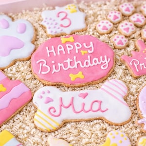 Puede incluir: Una colecci&oacute;n de galletas decoradas con tem&aacute;tica de perros en una caja rosa. Las galletas incluyen huellas de patas, una tarta de cumplea&ntilde;os, una galleta con forma de hueso con el nombre "Mia" y galletas con "Happy Birthday" y "Princess".