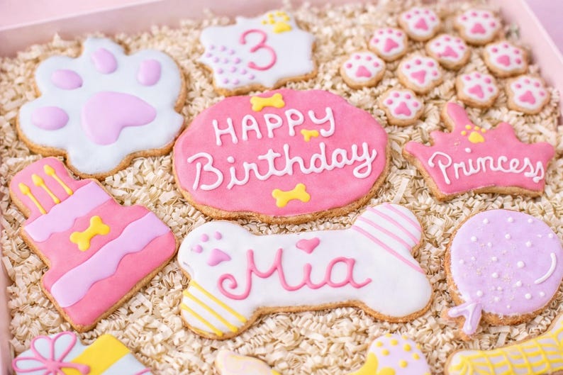 Puede incluir: Una colecci&oacute;n de galletas decoradas con tem&aacute;tica de perros en una caja rosa. Las galletas incluyen huellas de patas, una tarta de cumplea&ntilde;os, una galleta con forma de hueso con el nombre "Mia" y galletas con "Happy Birthday" y "Princess".