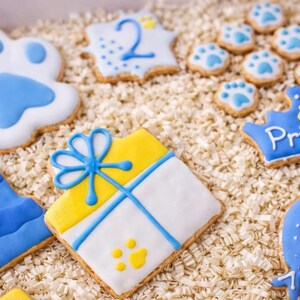 Puede incluir: Una variedad de galletas decoradas con glaseado azul y amarillo. Los dise&ntilde;os incluyen huellas de patas, una corona con la palabra "Prince" y una caja de regalo. Las galletas est&aacute;n dispuestas sobre un lecho de papel triturado.