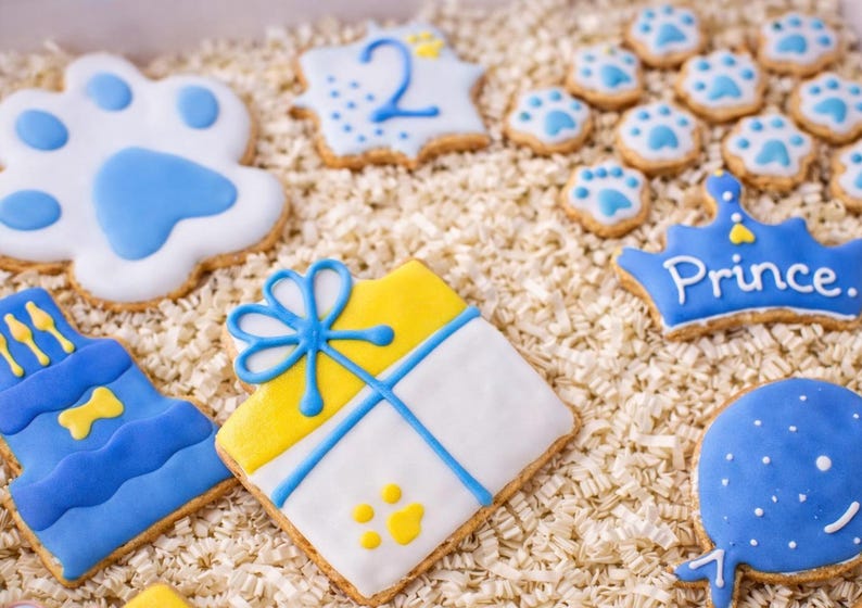 Puede incluir: Una variedad de galletas decoradas con glaseado azul y amarillo. Los dise&ntilde;os incluyen huellas de patas, una corona con la palabra "Prince" y una caja de regalo. Las galletas est&aacute;n dispuestas sobre un lecho de papel triturado.