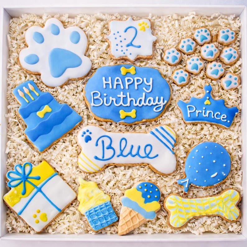 Puede incluir: Una caja de galletas decoradas con tem&aacute;tica de perros. Las galletas tienen varias formas, incluyendo huellas de patas, un hueso, un pastel, un regalo y conos de helado. Est&aacute;n decoradas con glaseado azul y amarillo y las palabras "Happy Birthday" y "Blue".