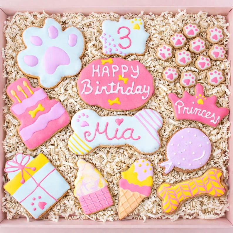 Puede incluir: Una caja de galletas decoradas para perros en una caja rosa. Las galletas tienen varias formas, incluyendo una huella de pata, el n&uacute;mero 3, un pastel, un hueso, una corona, un regalo, un helado y una piruleta. Las galletas est&aacute;n decoradas con glaseado rosa, blanco, amarillo y azul. Algunas galletas tienen las palabras "Happy Birthday" y "Mia".