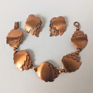 May include: A vintage copper-toned jewellery set comprising a bracelet and matching clip-on earrings. The bracelet and earrings are designed with layered leaf shapes, showcasing a warm, metallic finish. The set is displayed on a white surface.