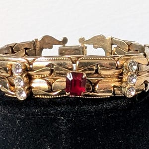 Peut inclure: Un bracelet à maillons en métal doré orné d'une pierre rouge rectangulaire centrale, flanquée de deux rangées verticales de trois strass ronds transparents, avec des pierres rouges rectangulaires aux extrémités et un fermoir en métal.