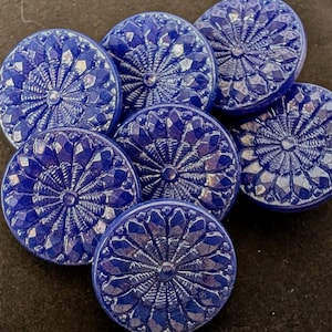 May include: Seven round, cobalt blue buttons with a detailed, radial design. Each button has a central point and faceted edges, giving a textured, jewel-like appearance. The buttons are arranged on a dark surface.