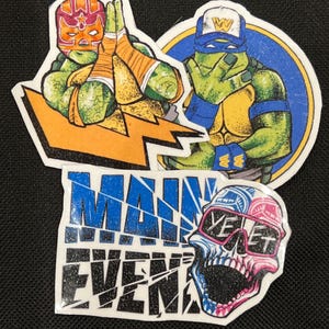 May include: Three colorful iron-on patches featuring Teenage Mutant Ninja Turtles characters. The first patch shows a green turtle wearing a red and orange wrestling mask with a lightning bolt behind him. The second patch shows a green turtle wearing a blue bandana and a yellow baseball cap with the letter W on it. The third patch shows a skull with sunglasses and the word "YET" on it. The patches are all on a black background and have a glittery finish.