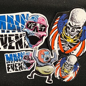 May include: Five colorful stickers featuring a skull with sunglasses, a skeleton with wings, a cartoon character, and the words "Main Event" in a broken font.