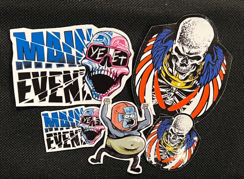 May include: Five colorful stickers featuring a skull with sunglasses, a skeleton with wings, a cartoon character, and the words "Main Event" in a broken font.