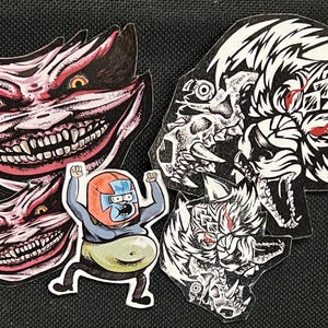 May include: Five stickers featuring different cartoon characters. The stickers are on a black background. One sticker features a white wolf with red eyes. Another sticker features a red and white creature with a large smile and sharp teeth. Another sticker features a red and white creature with a large smile and sharp teeth. Another sticker features a red and white creature with a large smile and sharp teeth. The last sticker features a cartoon character wearing a blue suit and a red helmet.