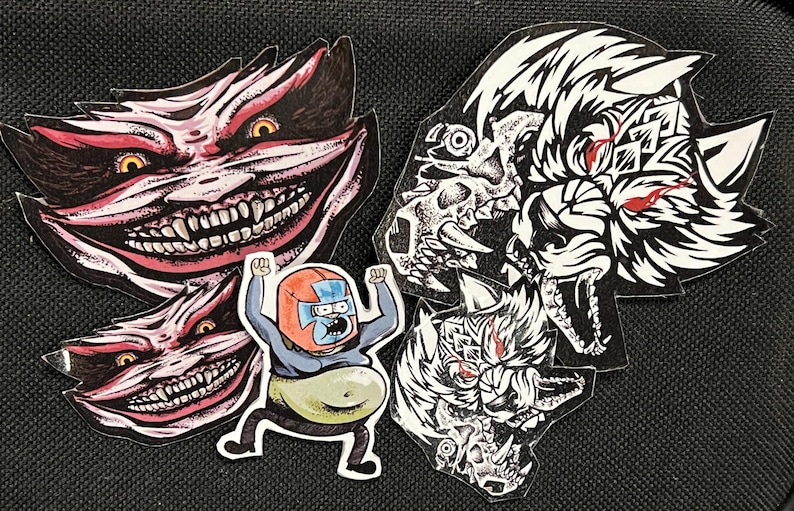 May include: Five stickers featuring different cartoon characters. The stickers are on a black background. One sticker features a white wolf with red eyes. Another sticker features a red and white creature with a large smile and sharp teeth. Another sticker features a red and white creature with a large smile and sharp teeth. Another sticker features a red and white creature with a large smile and sharp teeth. The last sticker features a cartoon character wearing a blue suit and a red helmet.