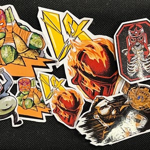 May include: A set of eight colorful stickers featuring wrestlers in various poses. The stickers are on a black background. The stickers are all different sizes and shapes. Some of the stickers have a lightning bolt design. One sticker features a wrestler with a skeleton inside.