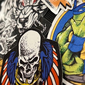 May include: A collection of colorful stickers featuring a skull with wings, a wolf, and a Teenage Mutant Ninja Turtle. The stickers are on a black background.