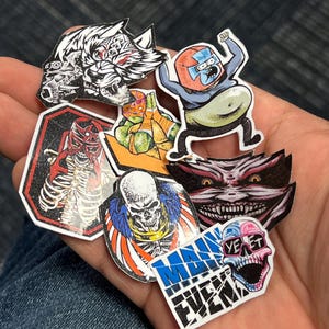 May include: A collection of colorful stickers featuring various characters and designs. The stickers include a wolf, a skeleton, a skull, a cartoon character, a Teenage Mutant Ninja Turtle, and a monster with sharp teeth. The stickers are on a blue denim background.