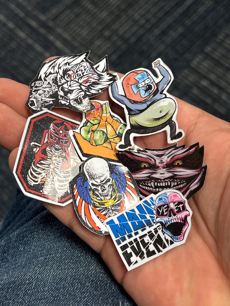 May include: A collection of colorful stickers featuring various characters and designs. The stickers include a wolf, a skeleton, a skull, a cartoon character, a Teenage Mutant Ninja Turtle, and a monster with sharp teeth. The stickers are on a blue denim background.